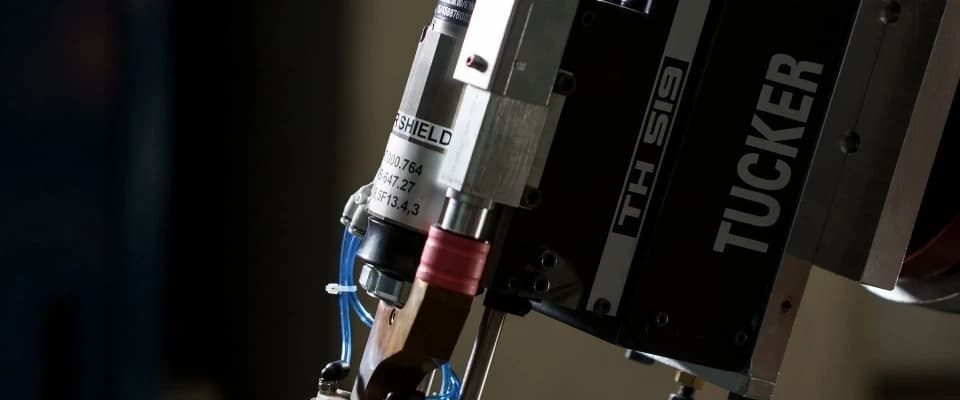 A close-up of a Tucker TH519 stud-welding head on a robot, black with the Tucker name down its side, its stud feed and shielding nozzle pointing down, against a dark background.